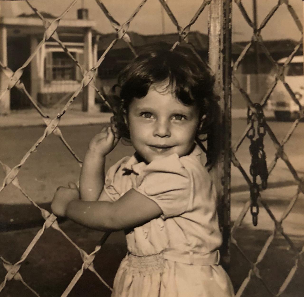 Uma fotografia antiga de uma menina de cerca de quatro anos. Ela tem os cabelos lisos e curtos e usa um vestido branco.